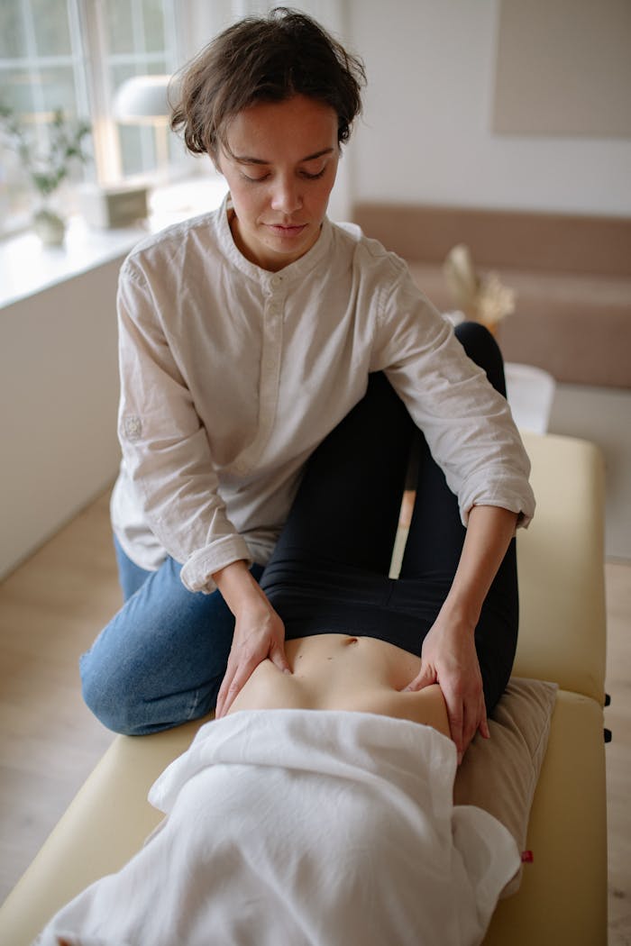A massage therapist performs a calming abdominal massage on a woman in a peaceful, sunlit room.